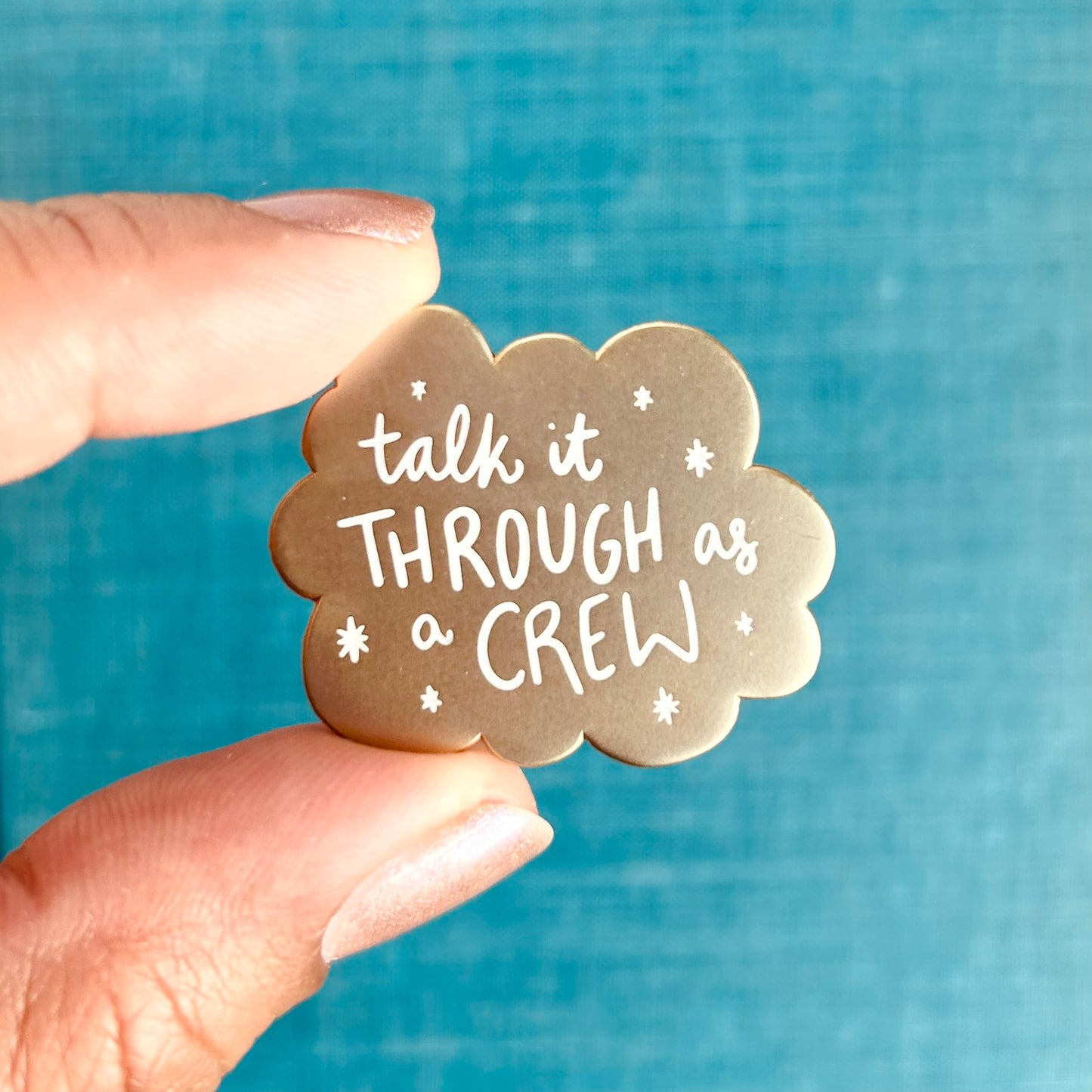 A small matte gold enamel pin with scalloped edges and white stars around the words "talk it through as a crew"