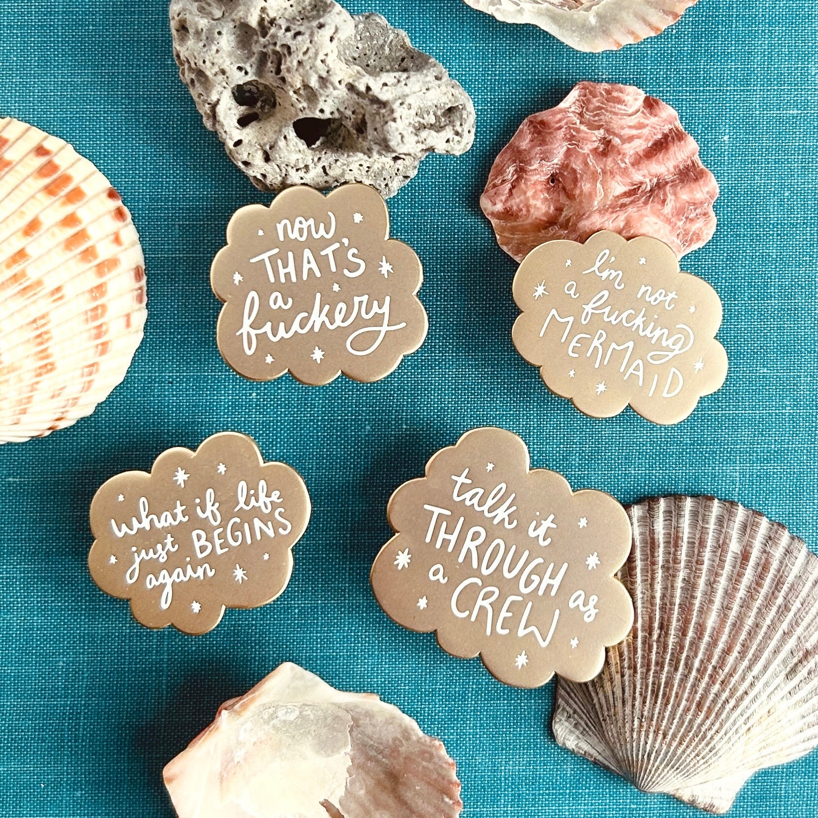 A collection of four gold scalloped enamel pins surrounded by shells