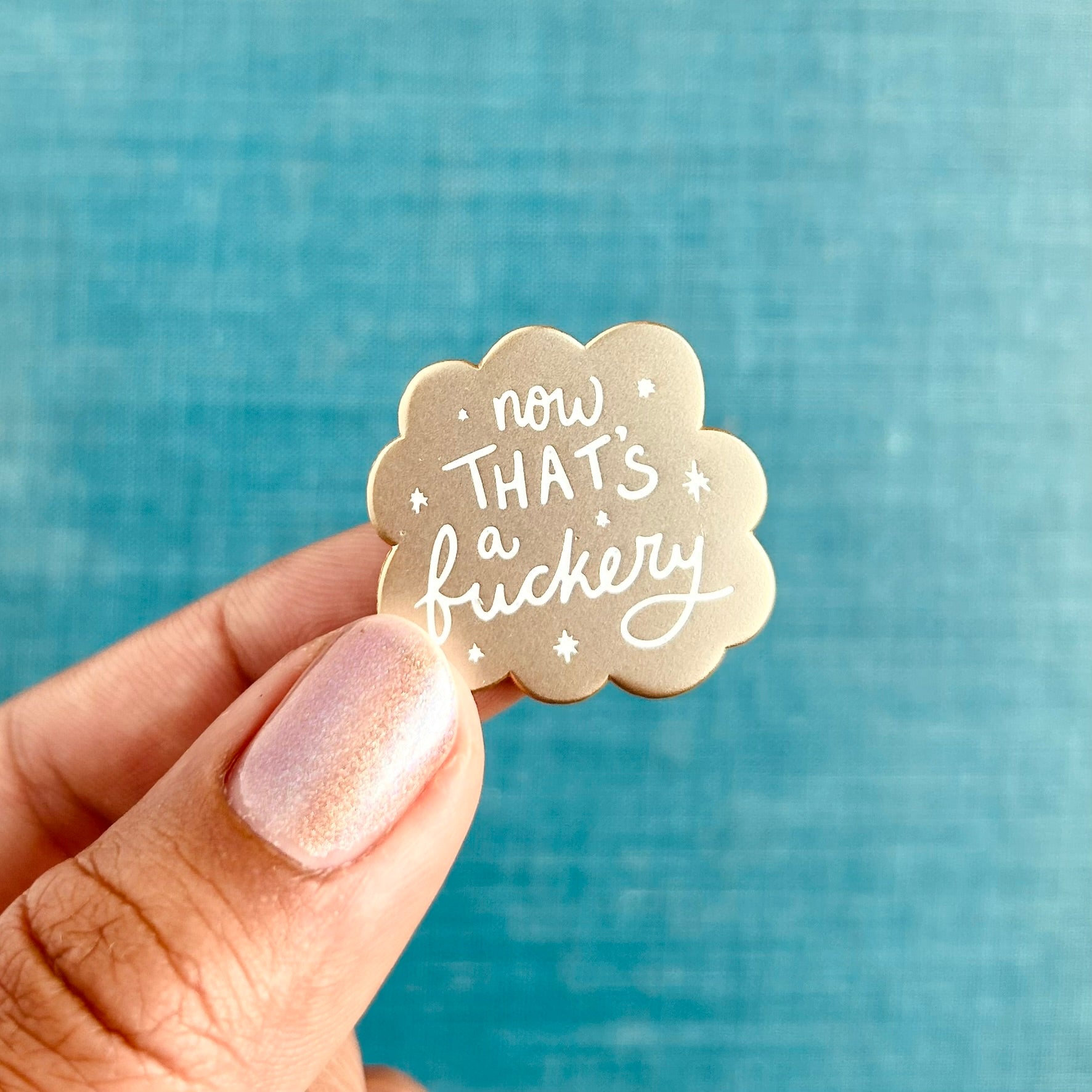 A small matte gold enamel pin with scalloped edges and white stars around the words "now that's a fuckery"