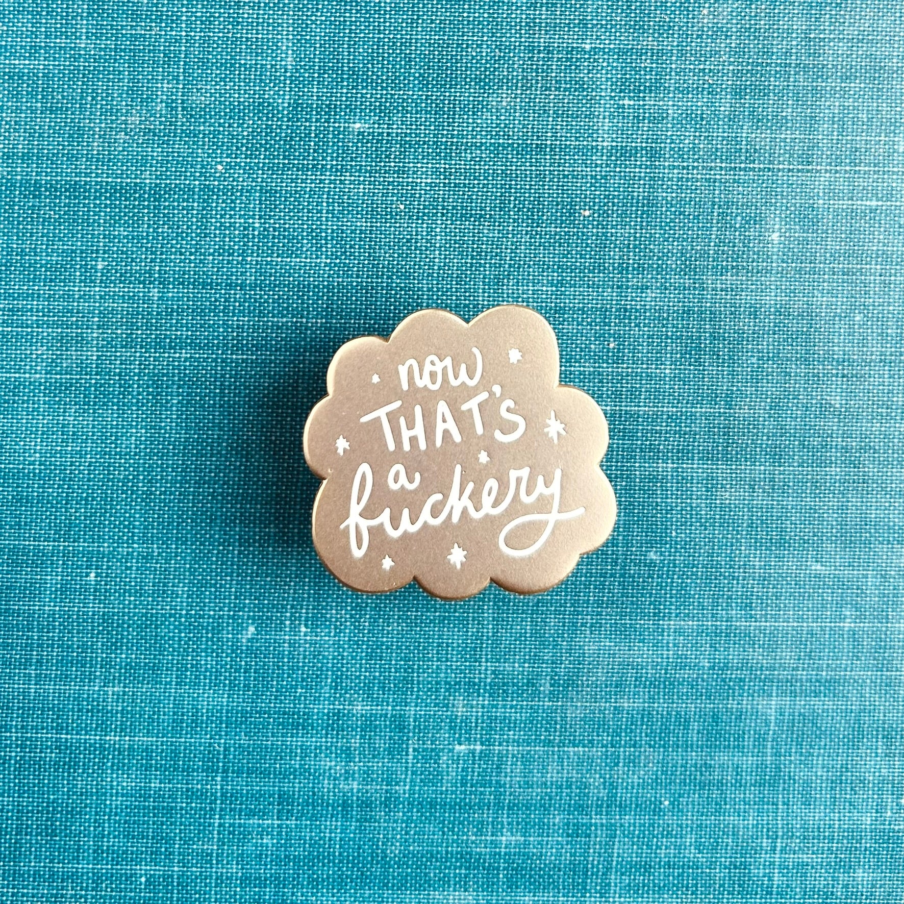 A small matte gold enamel pin with scalloped edges and white stars around the words "now that's a fuckery"