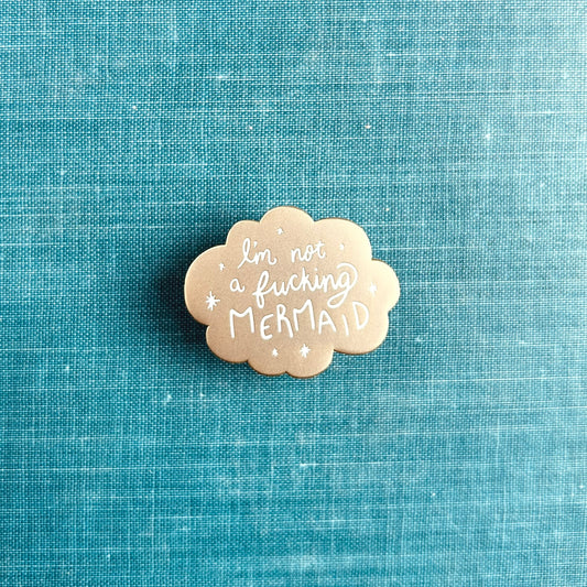 A small matte gold enamel pin with scalloped edges and white stars around the words "I'm not a fucking mermaid"