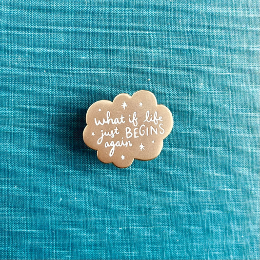 A small matte gold enamel pin with scalloped edges and white stars around the words "what if life just begins again?"