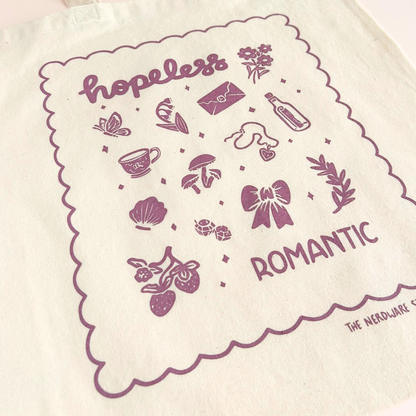 tote bag with illustrations in purple that say hopeless romantic with romantic flowers, a butterfly, mushrooms, strawberries, and a scalloped border