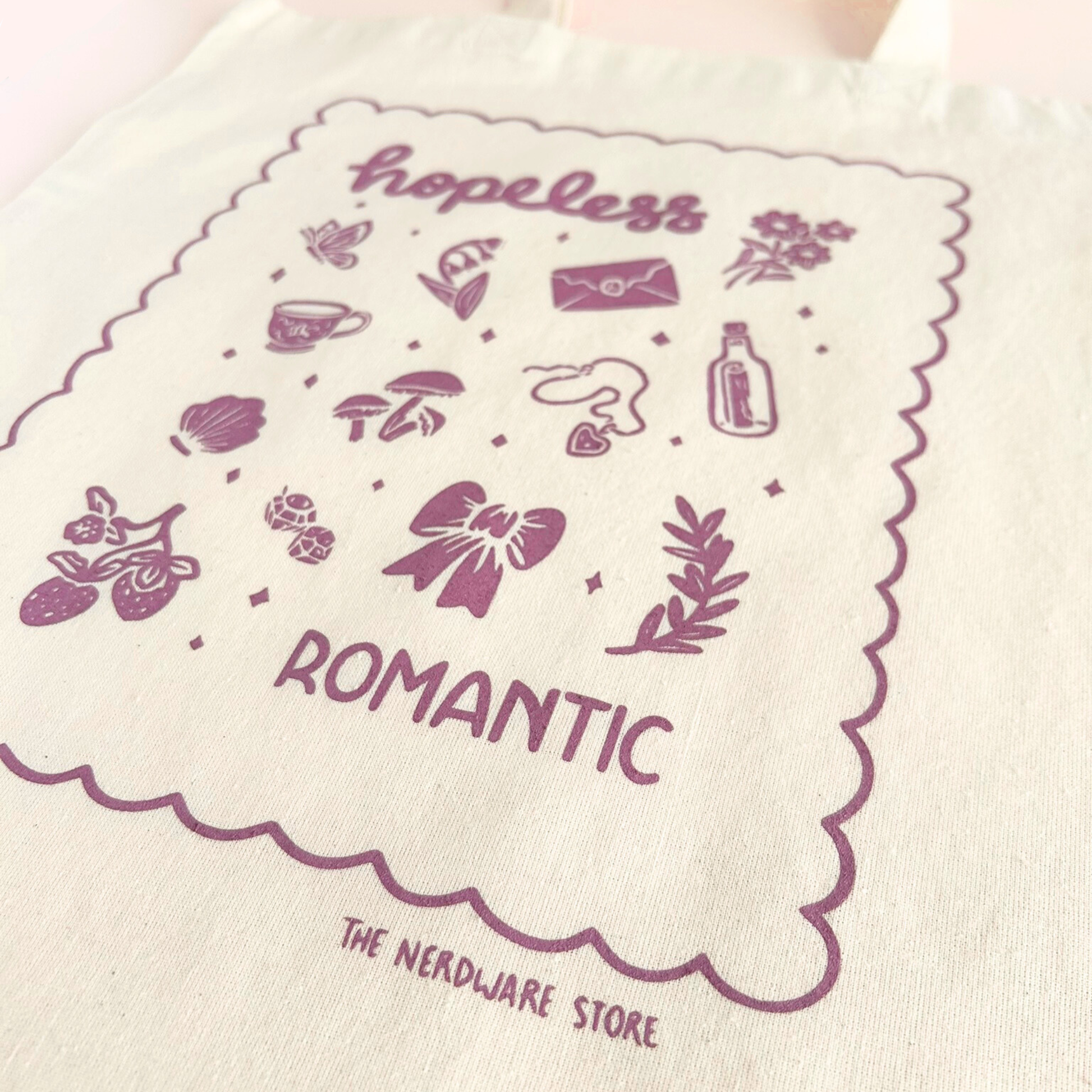 tote bag with illustrations in purple that say hopeless romantic with romantic flowers, a butterfly, mushrooms, strawberries, and a scalloped border