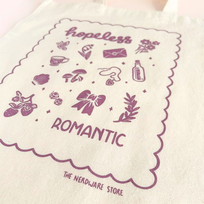 tote bag with illustrations in purple that say hopeless romantic with romantic flowers, a butterfly, mushrooms, strawberries, and a scalloped border