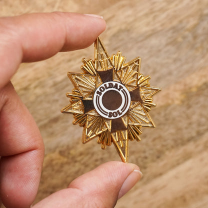 A gold star shaped enamel pin that says "soldat sol" on it.