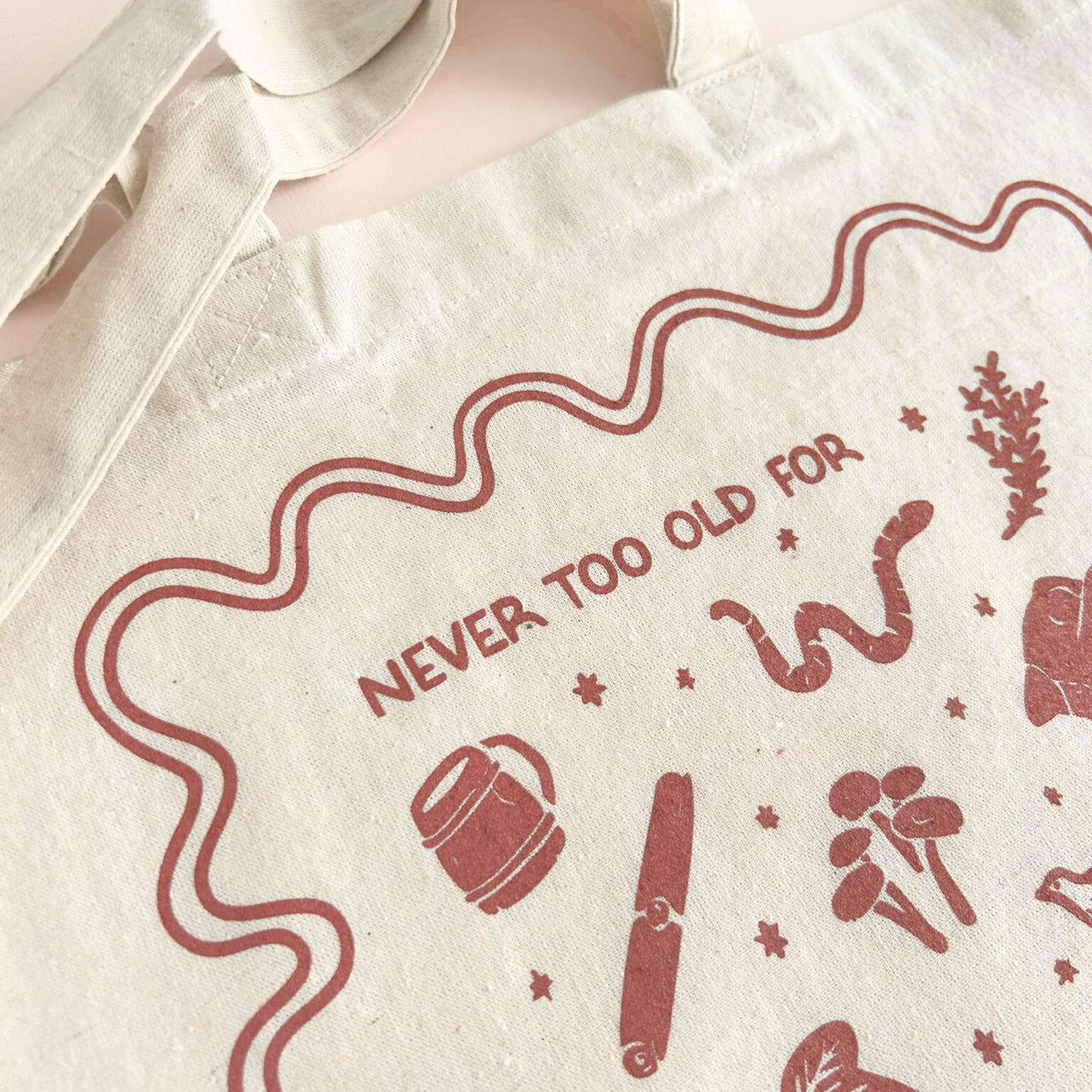 tote bag with designs in maroon that say never too old for stories with fantastical illustrations of a book, locket, moth, leaf, and more