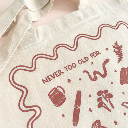tote bag with designs in maroon that say never too old for stories with fantastical illustrations of a book, locket, moth, leaf, and more