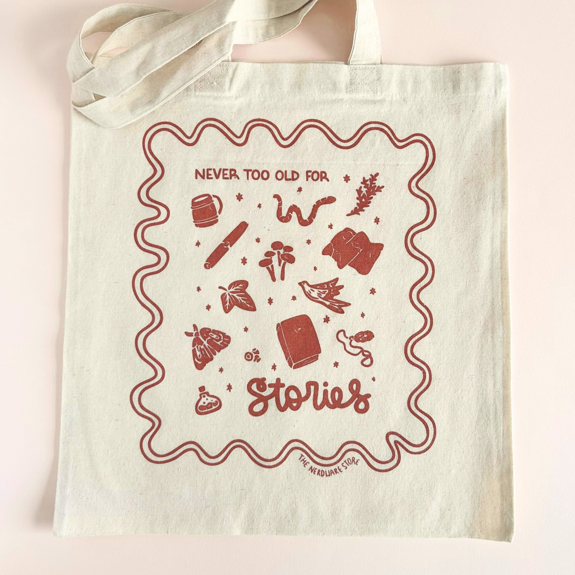 tote bag with designs in maroon that say never too old for stories with fantastical illustrations of a book, locket, moth, leaf, and more