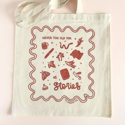 tote bag with designs in maroon that say never too old for stories with fantastical illustrations of a book, locket, moth, leaf, and more