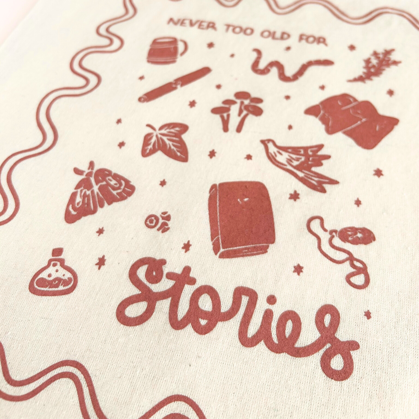 tote bag with designs in maroon that say never too old for stories with fantastical illustrations of a book, locket, moth, leaf, and more