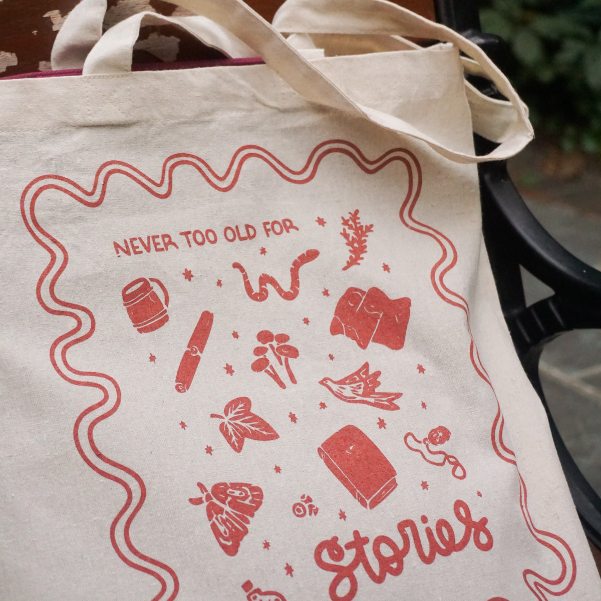 tote bag with designs in maroon that say never too old for stories with fantastical illustrations of a book, locket, moth, leaf, and more