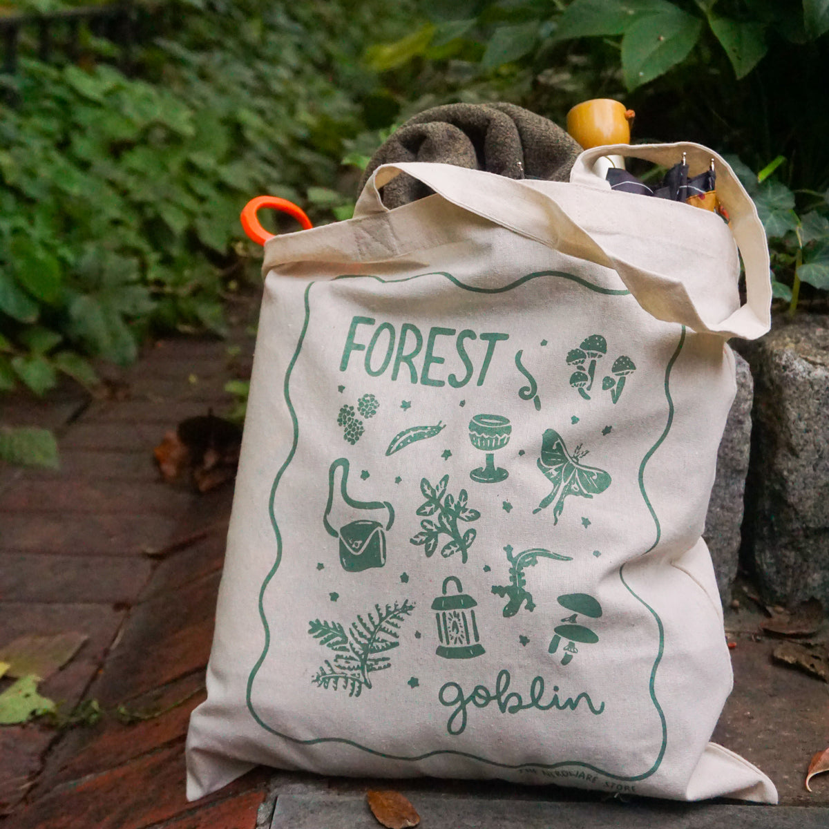 tote bag with designs in dark green that say forest goblin with fantastical illustrations of a slug, a goblet, a satchel, foliage, a lantern, mushrooms, and a luna moth