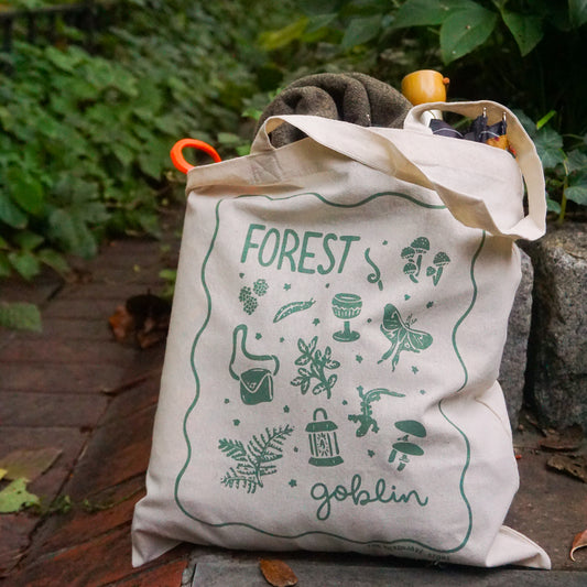 tote bag with designs in dark green that say forest goblin with fantastical illustrations of a slug, a goblet, a satchel, foliage, a lantern, mushrooms, and a luna moth