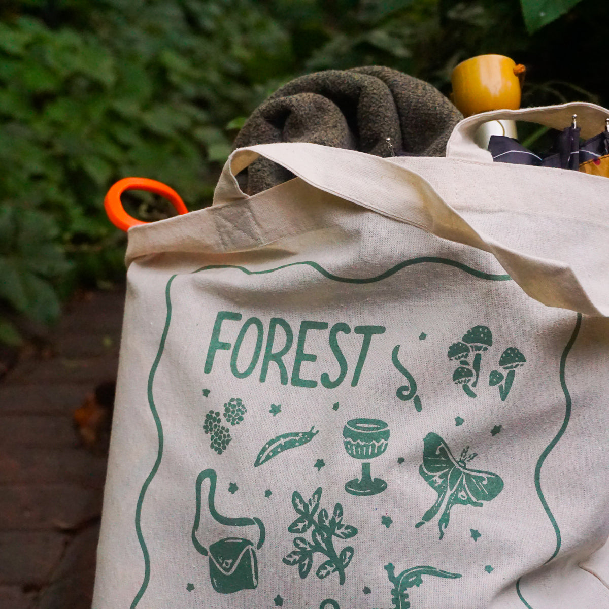 tote bag with designs in dark green that say forest goblin with fantastical illustrations of a slug, a goblet, a satchel, foliage, a lantern, mushrooms, and a luna moth