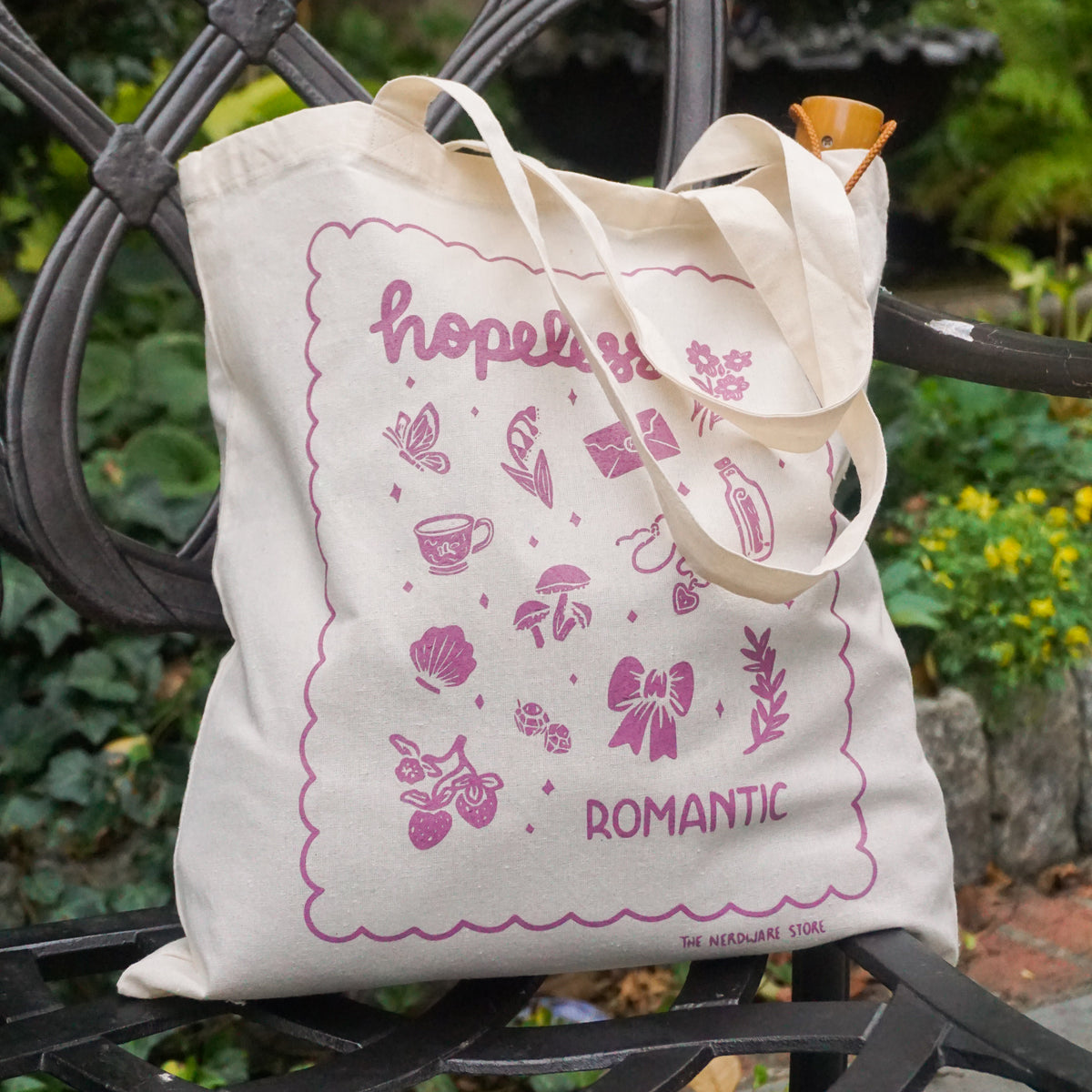 tote bag with illustrations in purple that say hopeless romantic with romantic flowers, a butterfly, mushrooms, strawberries, and a scalloped border