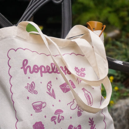 tote bag with illustrations in purple that say hopeless romantic with romantic flowers, a butterfly, mushrooms, strawberries, and a scalloped border