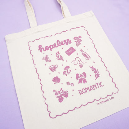 tote bag with illustrations in purple that say hopeless romantic with romantic flowers, a butterfly, mushrooms, strawberries, and a scalloped border