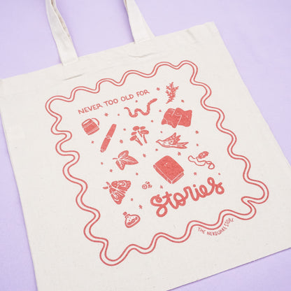 tote bag with designs in maroon that say never too old for stories with fantastical illustrations of a book, locket, moth, leaf, and more