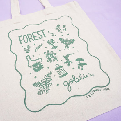 tote bag with designs in dark green that say forest goblin with fantastical illustrations of a slug, a goblet, a satchel, foliage, a lantern, mushrooms, and a luna moth