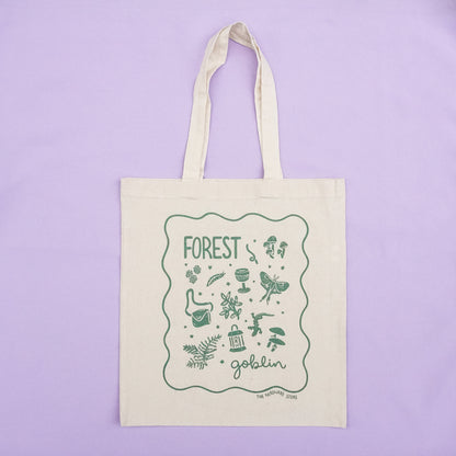 tote bag with designs in dark green that say forest goblin with fantastical illustrations of a slug, a goblet, a satchel, foliage, a lantern, mushrooms, and a luna moth