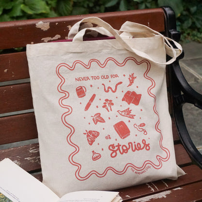 tote bag with designs in maroon that say never too old for stories with fantastical illustrations of a book, locket, moth, leaf, and more