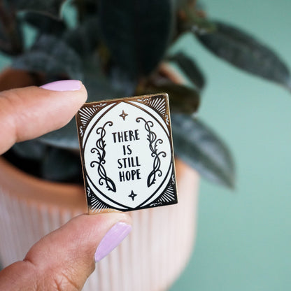 A hand holds up a small gold and light blue enamel pin with elvish scroll designns on it and the words "there is still hope."