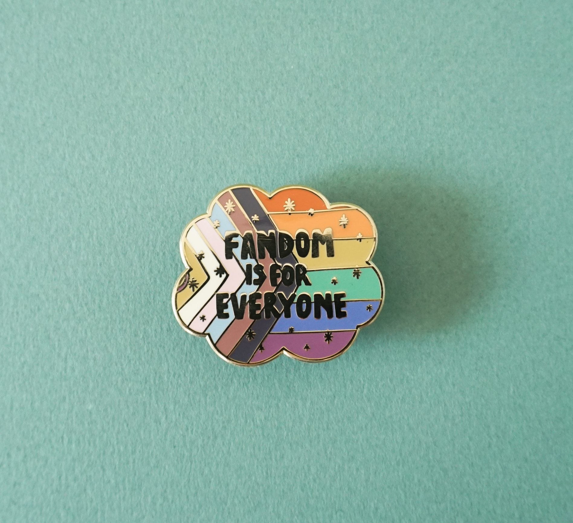 A gold enamel lapel pin showing the LGBTQIA+ pride flag with the words "Fandom is for everyone" over top.