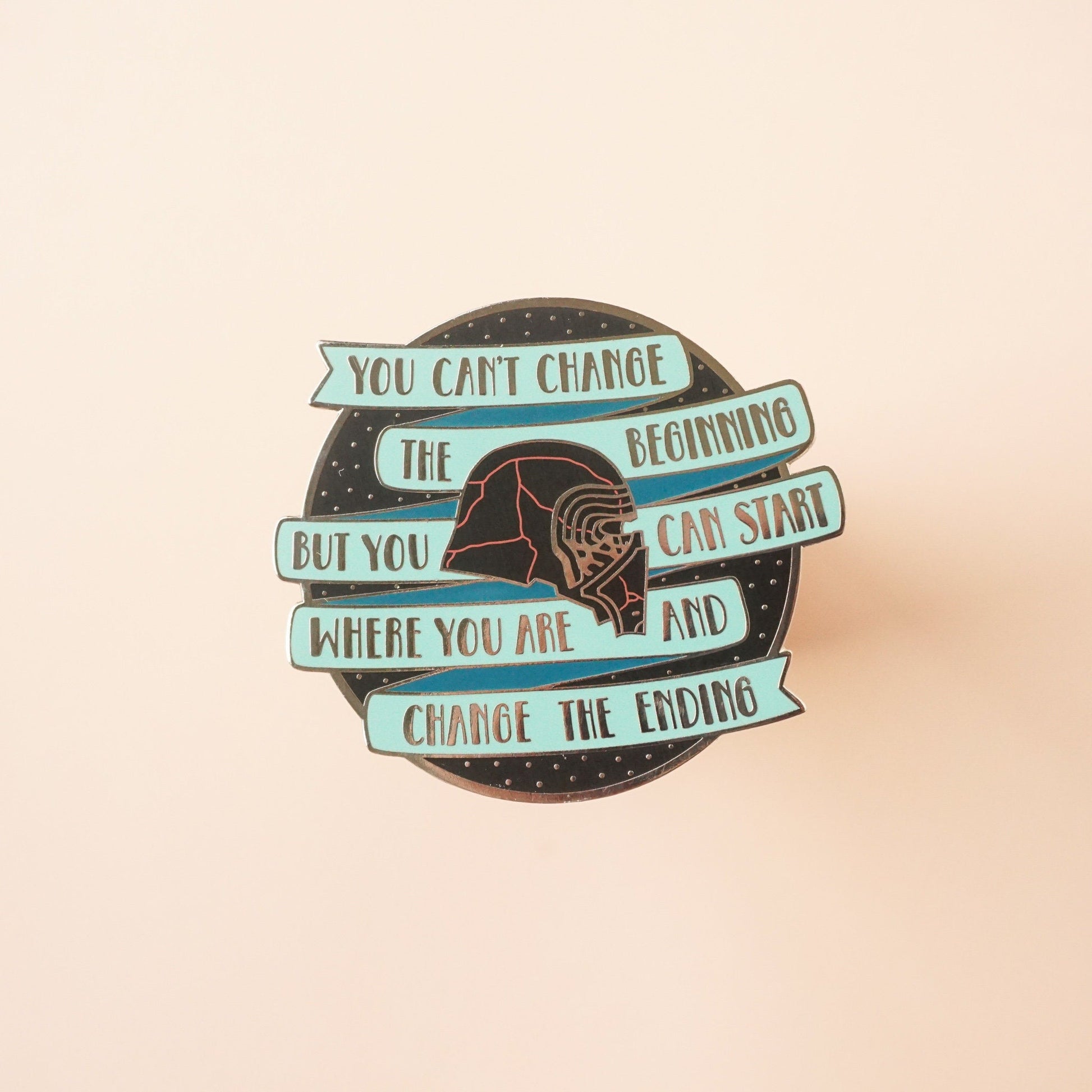 A round black pin with a teal banner on it that says "you cannot change the beginning, but you can start where you are and changing the ending" around kylo ren's broken helmet.