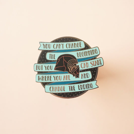 A round black pin with a teal banner on it that says "you cannot change the beginning, but you can start where you are and changing the ending" around kylo ren's broken helmet.