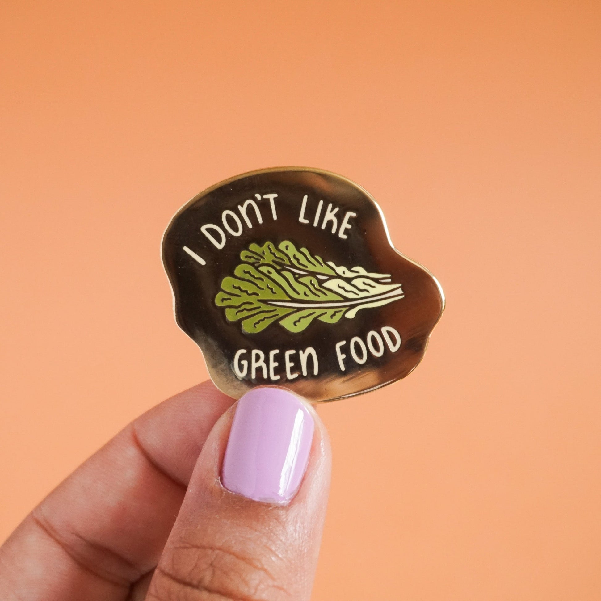 A small gold enamel pin that says "I don't like green food" with a romaine lettuce leaf in the center.