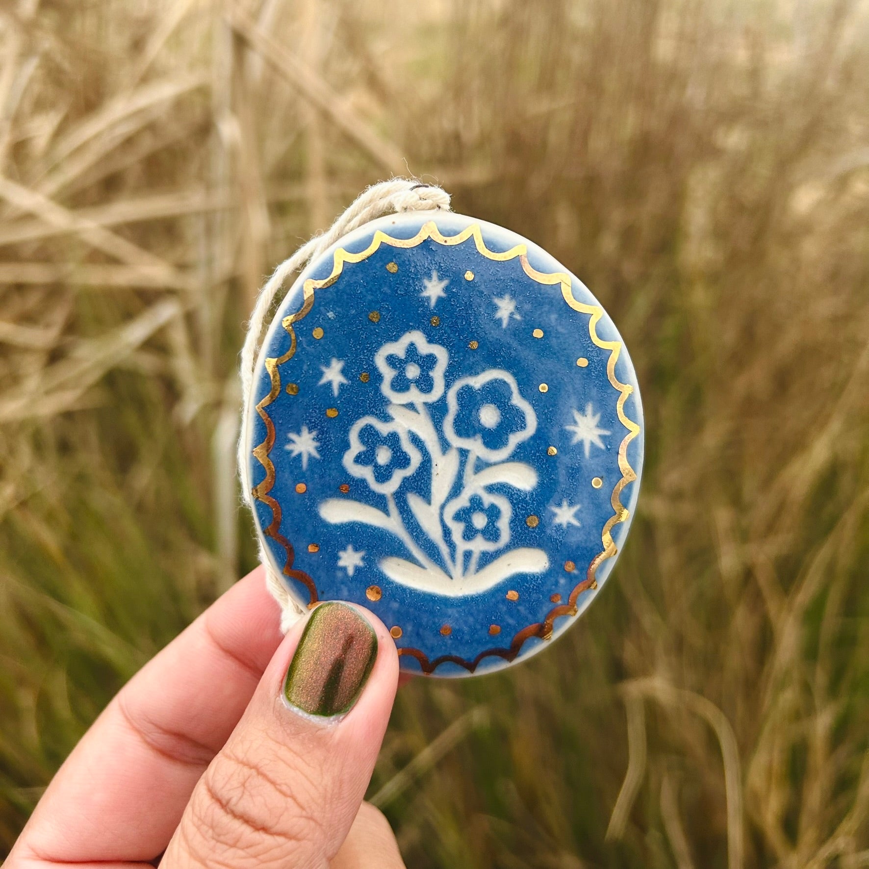 a hand holds up a bright blue ornament with some vintage style flowers carved into it and gold scalloped edges and gold dots
