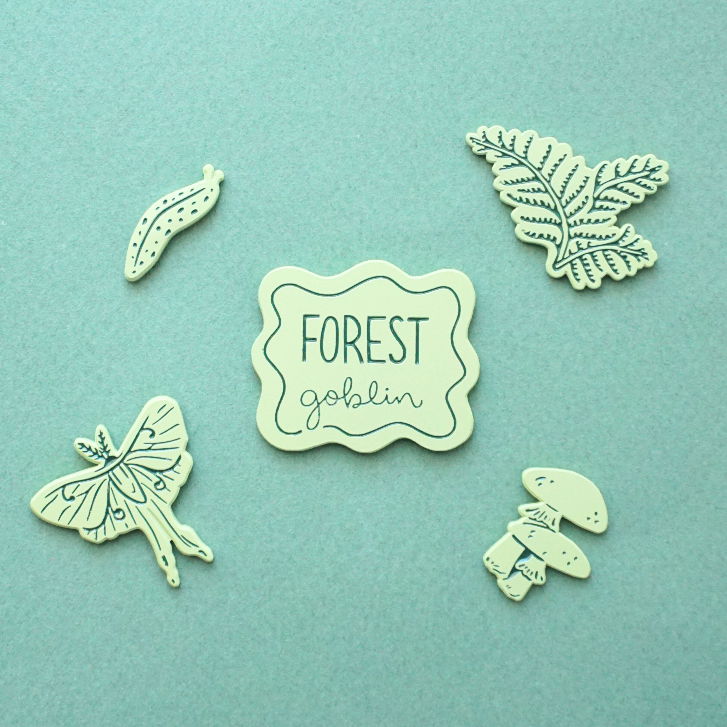 five light green pins of a slug, a luna moth, a fern, mushrooms, and a badge that says "forest goblin"