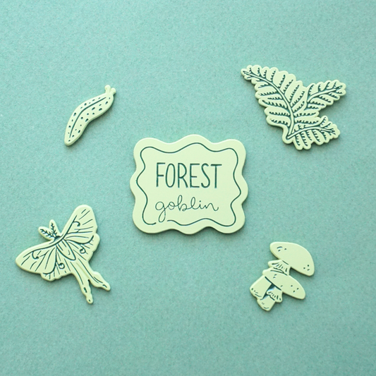 five light green pins of a slug, a luna moth, a fern, mushrooms, and a badge that says "forest goblin"