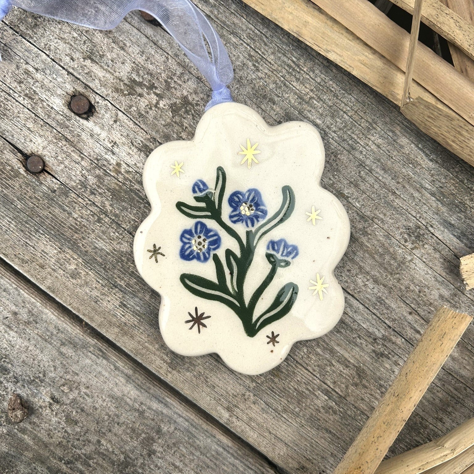 A scallop edged white ceramic ornament with blue forget me not blossoms and green leaves surrounded by gold stars and with a blue ribbon on the ornament.