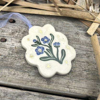 A scallop edged white ceramic ornament with blue forget me not blossoms and green leaves surrounded by gold stars and with a blue ribbon on the ornament.