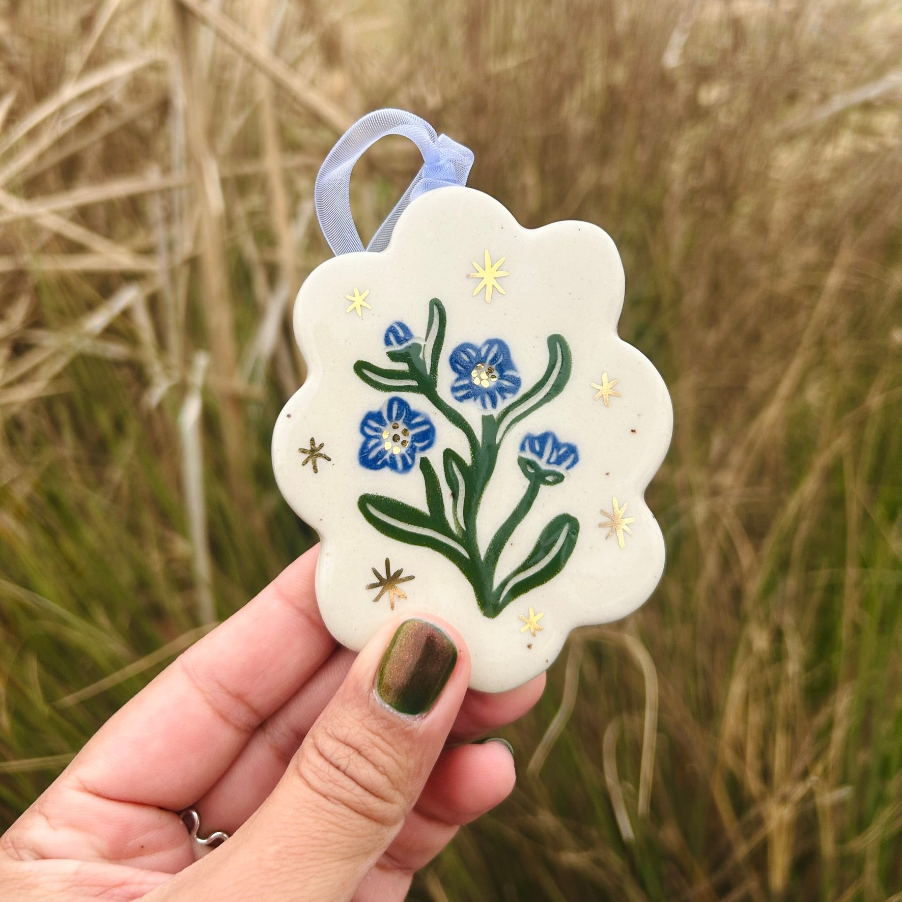 A hand holds up a scallop edged white ceramic ornament with blue forget me not blossoms and green leaves surrounded by gold stars and with a blue ribbon on the ornament and reeds in the background of the photo.