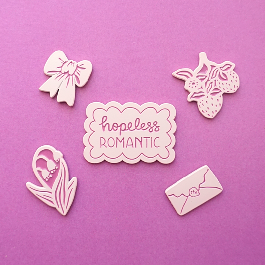 five light pink enamel pins of a bow, strawberries, lillies of the valley, an envelope, and the words "hopeless romantic"