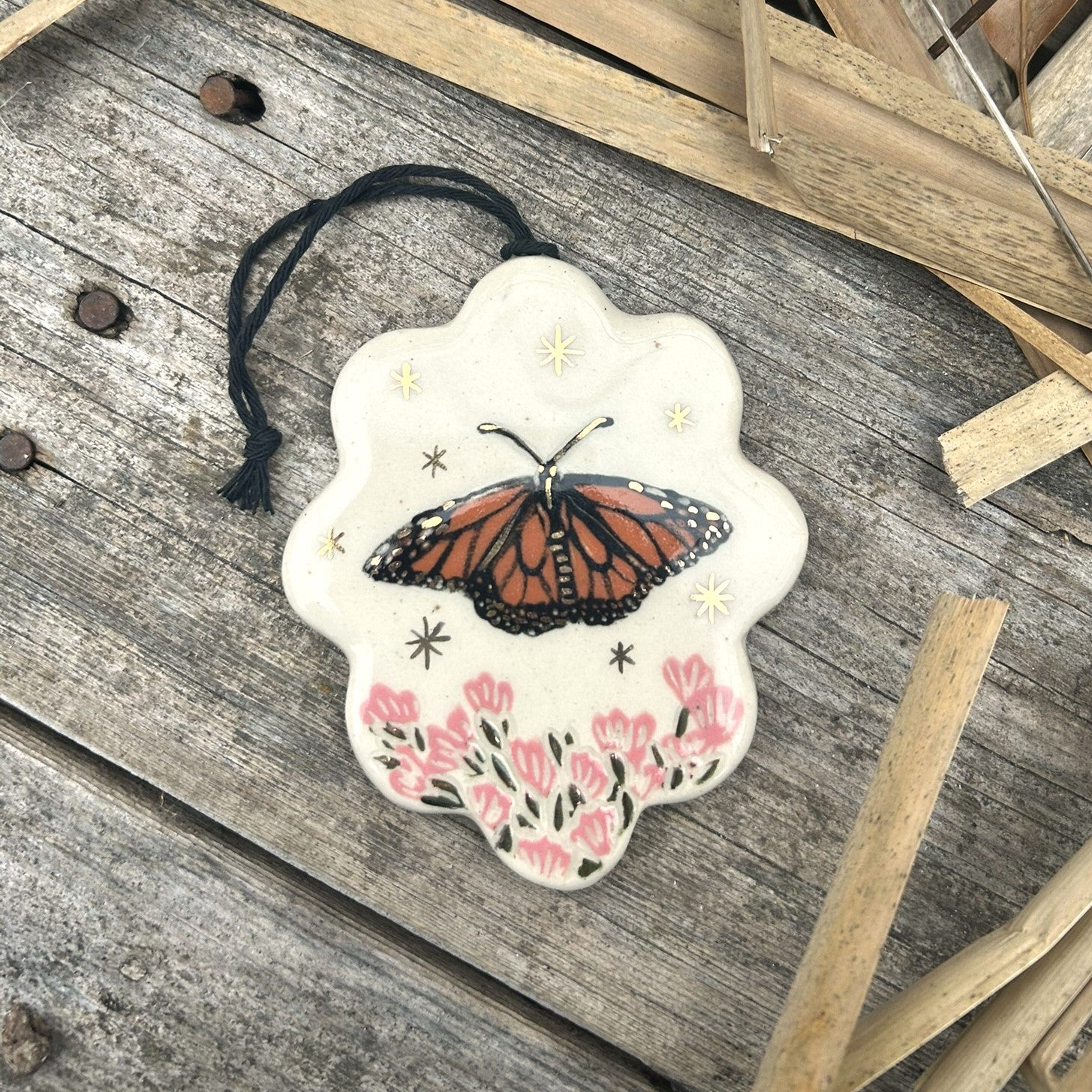 a scallop edged ornament with a monarch butterfly on it and pink flowers below and gold stars surrounding it.