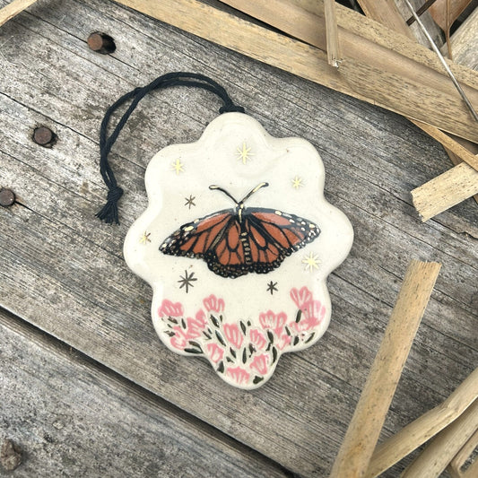 a scallop edged ornament with a monarch butterfly on it and pink flowers below and gold stars surrounding it.