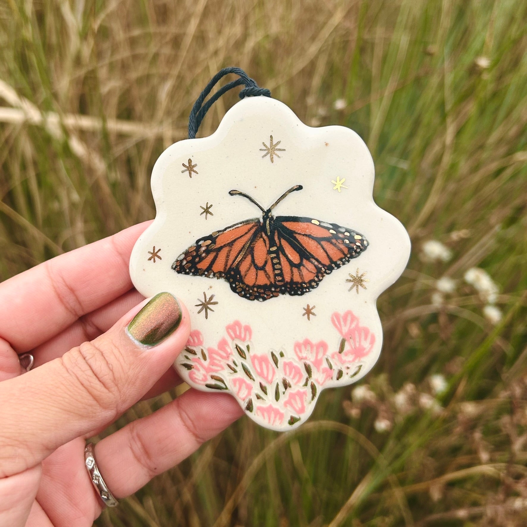 A hand holds up a scallop edged ornament with a monarch butterfly on it and pink flowers below and gold stars surrounding it. The background shows green and brown reeds.