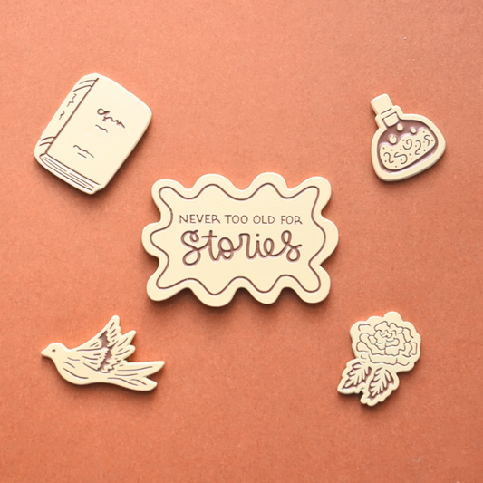 five light peach enamel pins of a book, a magic potion, a dove, and a rose and the words "never too old for stories."